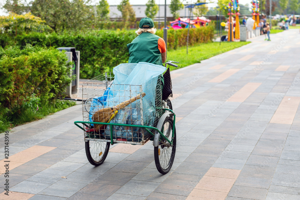 Fototapeta premium Urban street cleaner riding bicycle with garbage bag through municipal park on sunny day. Sweeper worker with bicycle on pavement at city street