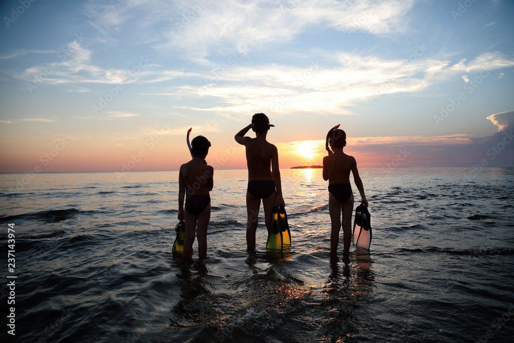 Silhouette-Rear View Of Three Boys Standing In The Sea With Diving Equipment And Looking At Horizon On Beautiful Sunset
