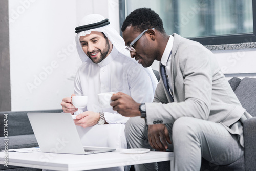 Multicultural businessmen holding cups and looking at laptop in modern office