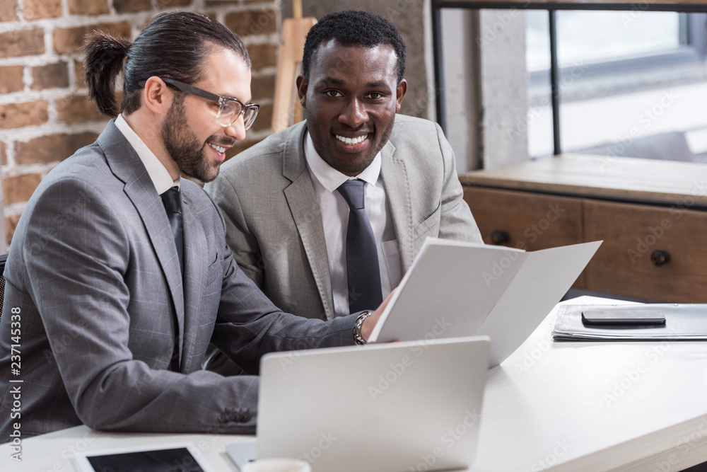 handsome businessman reading document while african american partner looking at camera in office handsome businessman reading document while african american partner looking at camera in office