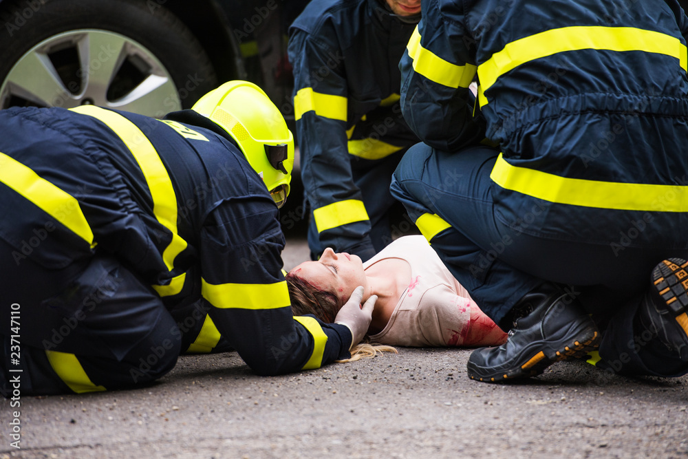 Three firefighters helping a young injured woman lying on the road after an accident.
