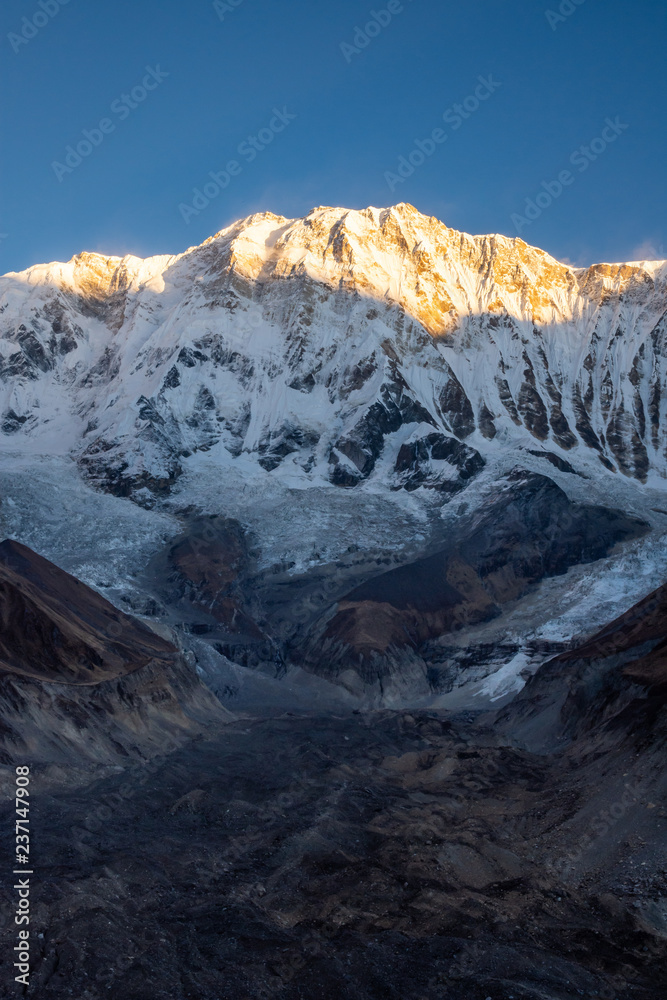 Vertical photo of Annapurna 1 and its glacier morraine during sunrise (golden hour) against blue sky, Himalayas