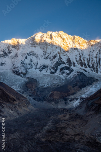 Vertical photo of Annapurna 1 and its glacier morraine during sunrise (golden hour) against blue sky, Himalayas