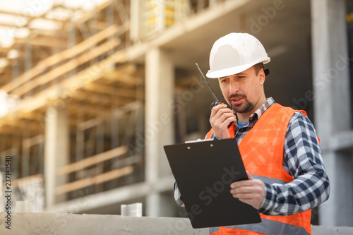 Developing engineer wearing white safety vest and hardhat with walkie talkie and clipboard inspecting construction site photo