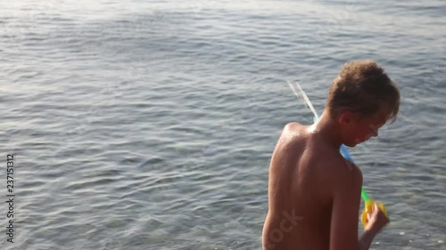 Back view of little cute kid playing gun water toy isolated on blue sea water background. Boy holding plastic pump shooting. Happy summer beach holiday concept. Real time full hd video footage. photo