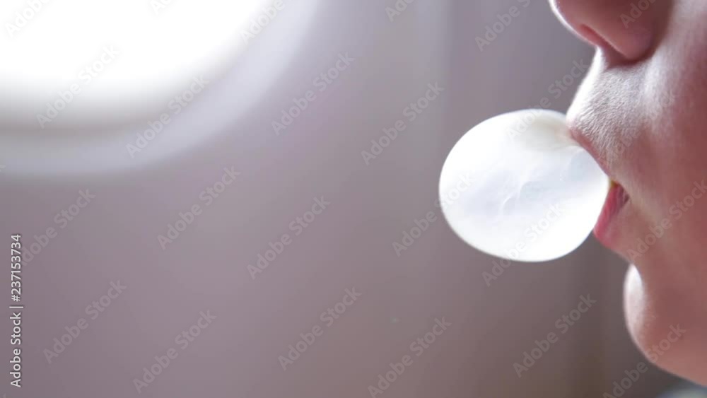 Closeup side view of cute little white kid blowing bubble chewing gum while sitting in his seat on board of plane. Real time full hd video footage.