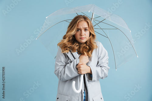 Image of displeased woman 20s wearing raincoat standing under transparent umbrella, isolated over blue background