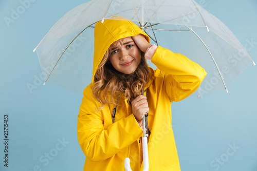 Image of smiling woman 20s wearing yellow raincoat standing under transparent umbrella, isolated over blue background