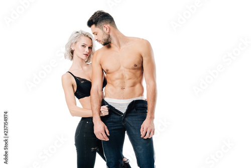 seductive woman looking at camera and unzipping pants of muscular man isolated on white