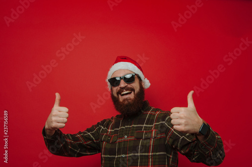 Cheerful bearded hipster man wearing santa claus hat and showing thumbs up over red background