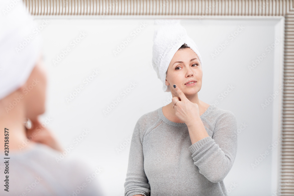 Middle aged caucasian woman with bath towel on the head looking at reflection in mirror in the morning, admires herself and smiling Middle aged caucasian woman with bath towel on the head looking at reflection in mirror in the morning, admires herself and smiling