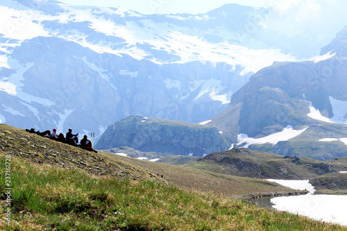 Group of four hikers is taking the rest on the slope and looking on high mountains, snow and frozen lake of Italian Alps