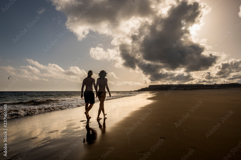 Naklejka premium young beautiful thin couple walking together with love viewed from rear on the shore with sunset and warm golden sky in backgrounds - friendship and relationship concept for people on vacation