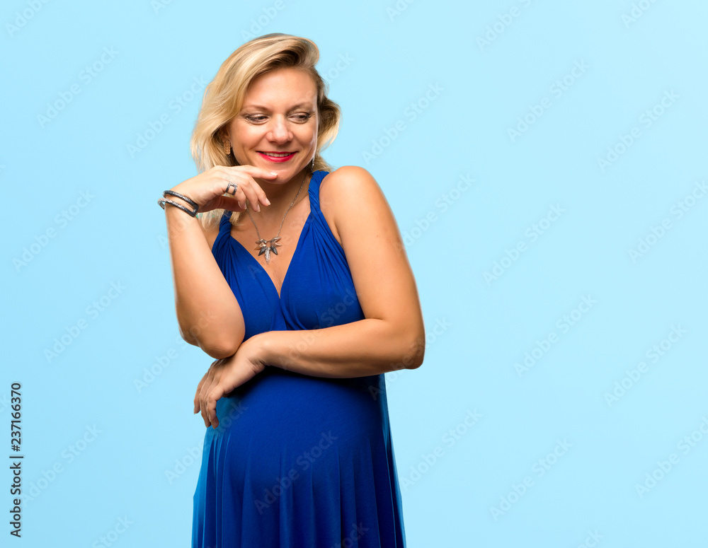 Pregnant blonde woman with blue dress laughing on isolated blue background Pregnant blonde woman with blue dress laughing on isolated blue background