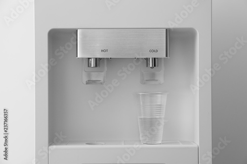 Modern water cooler with plastic cup on light background