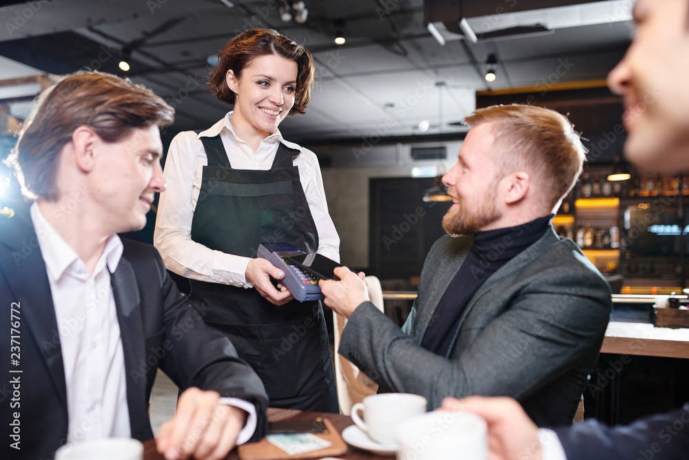 Smiling pretty young waitress in black apron giving terminal for payment to businessman in modern restaurant