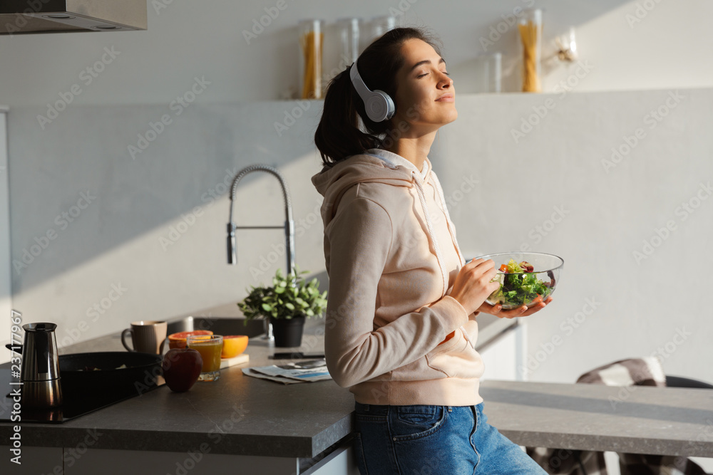 Happy healthy woman in the kitchen standing daily morning routine eat salad listening music in headphones. - obrazy, fototapety, plakaty 