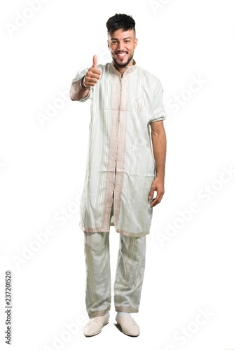 A full-length shot of a Arabic young man wearing typical arab clothes giving a thumbs up gesture and smiling on isolated white background