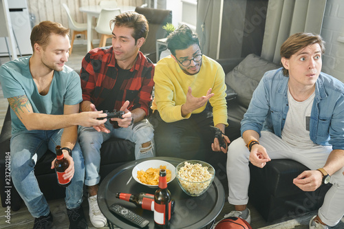 Young men resting on sofa with popcorn and alcohol drinks, they playing video games in turn