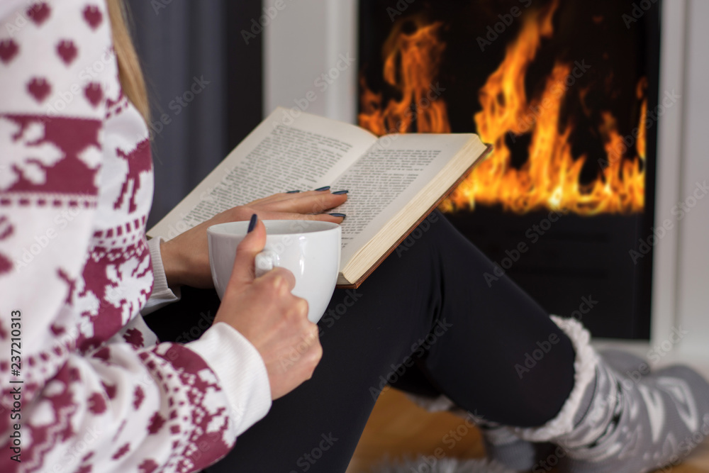 Young girl sitting in front of the fireplace and reading book and drinking  hot tea. Girl