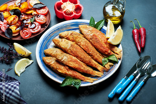 Fried seafood (‘red mullet’, ‘barbule’) on on a blue plate, dark background. Next plate with vegetable salad and red pepper photo