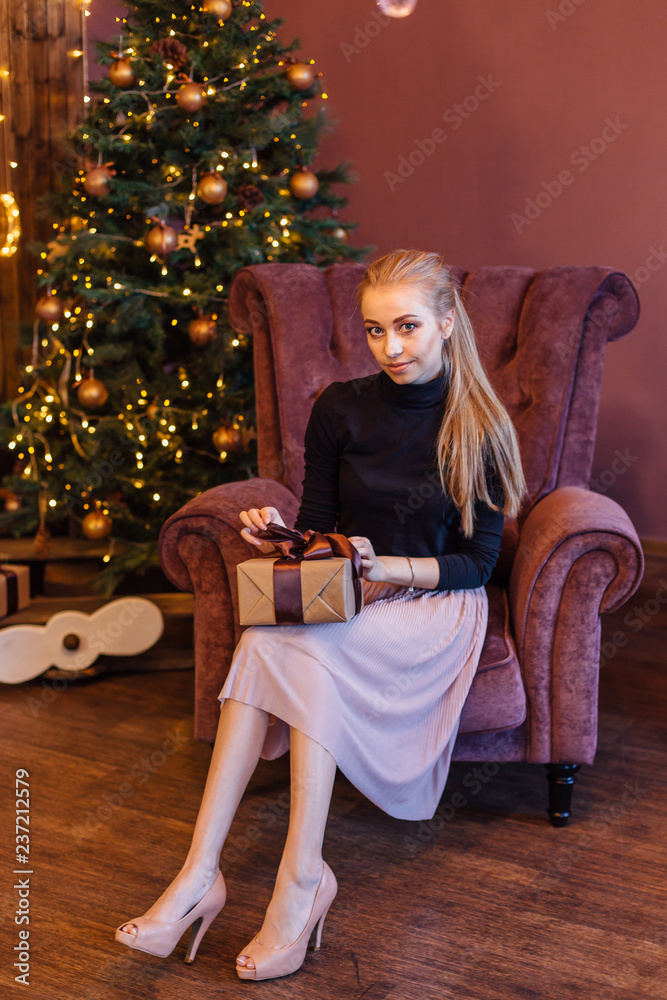 Beautiful young woman dressed in a sweater and skirt sitting on the arm chair next to the christmas tree with gift box in hands. Beautiful young woman dressed in a sweater and skirt sitting on the arm chair next to the christmas tree with gift box in hands.