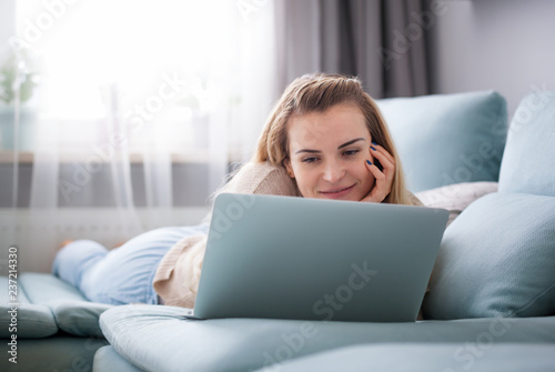 Smiling woman looking at laptop screen while lying on sofa at home, relaxing and resting