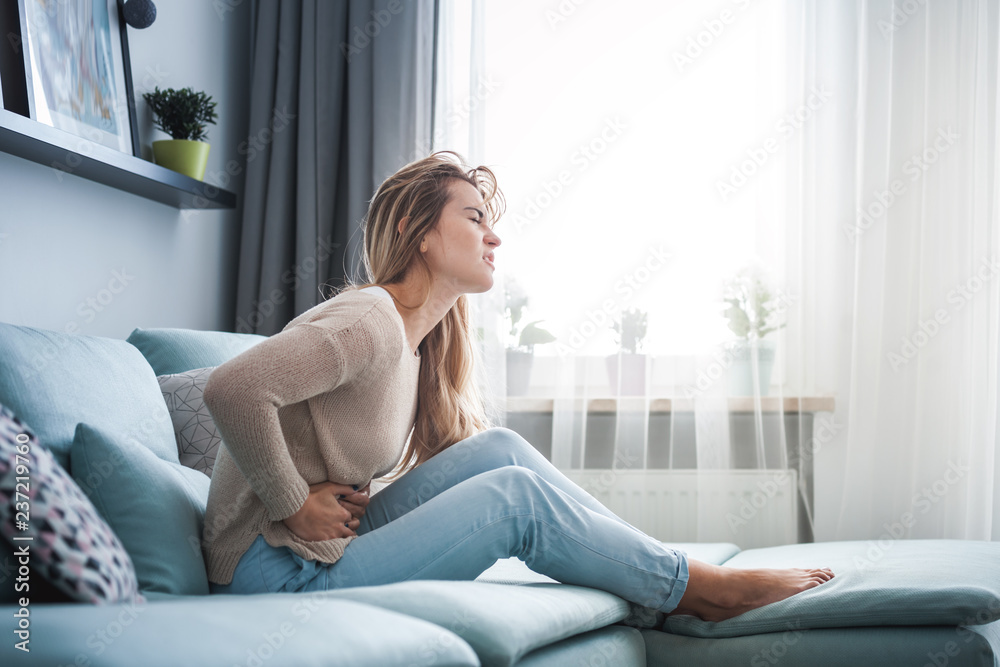 Young woman suffering from strong abdominal pain while sitting on sofa at home