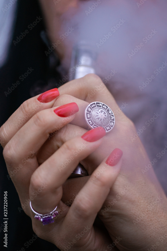 Caucasian woman with red nails manicure and antique ring on finger holds small vape. Smoking alternative vay. Woman exhales thick smoke. Life without cigarettes. Woman-vaper. Small e-cigarette. Caucasian woman with red nails manicure and antique ring on finger holds small vape. Smoking alternative vay. Woman exhales thick smoke. Life without cigarettes. Woman-vaper. Small e-cigarette.