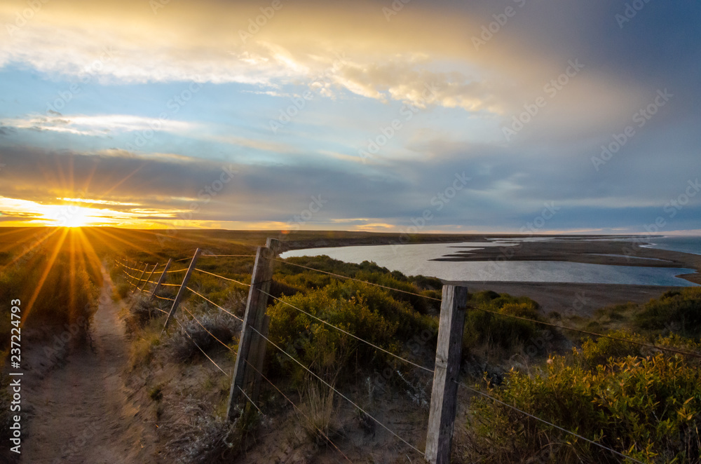 Landscape with a lake and a road with a wire fence on sunset with a golden and blue sky and sun rays Landscape with a lake and a road with a wire fence on sunset with a golden and blue sky and sun rays
