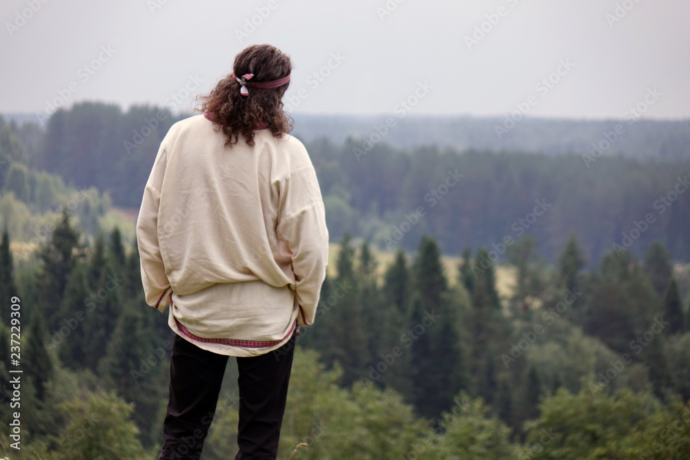 Naklejka premium A young man in a traditional folk costume in nature, looking into the distance. Green pine forest.