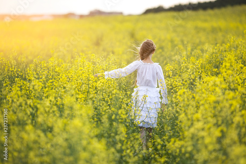A girl in a white dress is standing on a flowering rapeseed field on a sunny day.
