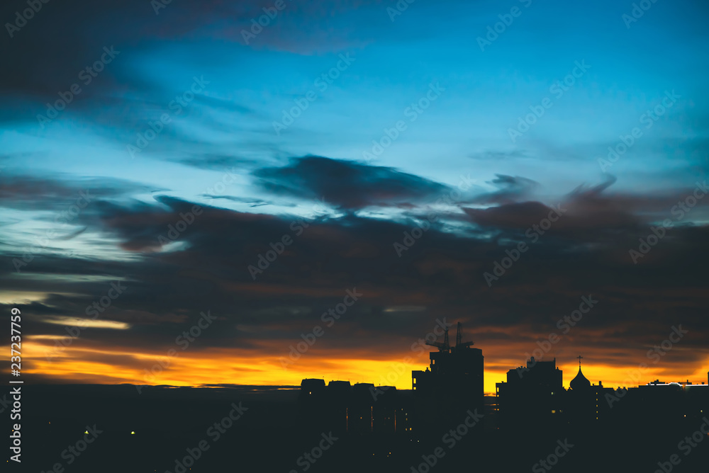 Cityscape with wonderful varicolored vivid dawn. Amazing dramatic blue cloudy sky above dark silhouettes of city buildings. Atmospheric background of orange sunrise in overcast weather. Copy space.