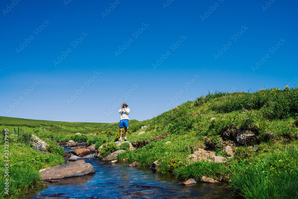 Traveler with camera on stone in mountain creek. Adventure of tourist. Hiking in mountains. Rich vegetation of highlands. Stream of clear water in brook. Vivid sunny landscape of majestic nature. Traveler with camera on stone in mountain creek. Adventure of tourist. Hiking in mountains. Rich vegetation of highlands. Stream of clear water in brook. Vivid sunny landscape of majestic nature.