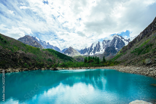 Mountain lake is surrounded by large stones and boulders on front of giant beautiful glacier. Amazing mountain in shape of pyramid. Snowy ridge under cloudy sky. Wonderful atmospheric landscape.