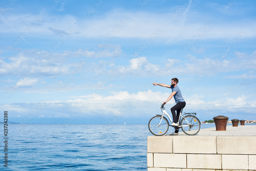 Fototapeta premium Young bearded man on bicycle on high paved stone sidewalk pointing at distant mountain on opposite shore on clear blue sea water and small sailing boat background. Active holiday and tourism concept.