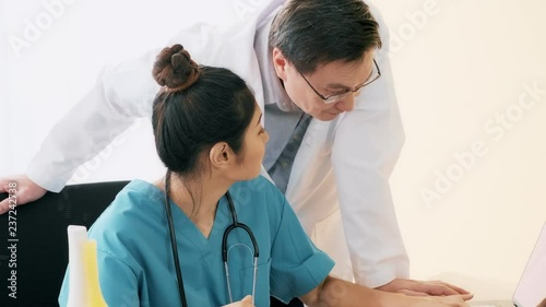 Senior asian man medical doctor harassing young medical doctor woman sitting on her desk. Young medical doctor office harassment concept.