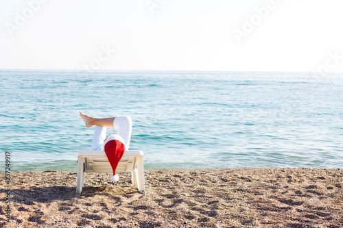 Woman in Christmas cap and white trousers lying on the beach chaise. Santa Claus on vacation. Christmas holiday. Winter resort. Christmas in Australia. 