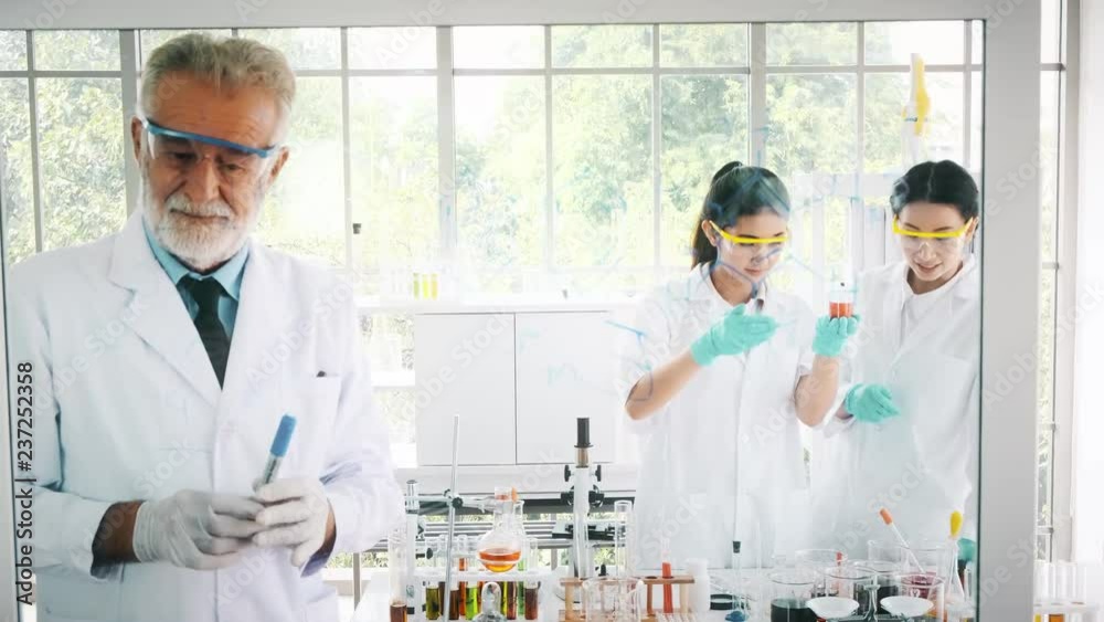 Group of chemists working in a lab. Young asian female chemists with senior caucasian chemist working together in lab, writing on glass display. Science concept. Group of chemists working in a lab. Young asian female chemists with senior caucasian chemist working together in lab, writing on glass display. Science concept.