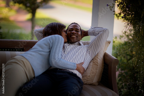Mid-adult woman hugging and kissing her husband as he relaxes on a couch.
