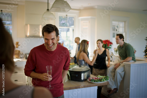 Laughing young man holding a champagne flute while inside a kitchen with friends. photo