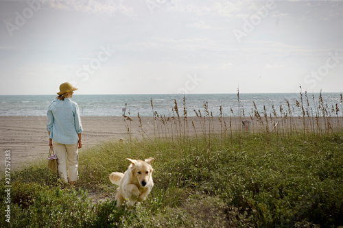 Mid-adult woman standing with a dog on grassy dunes of a beach.