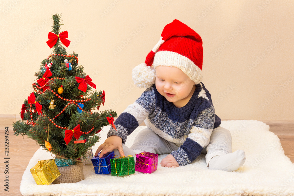 Funny baby in Santa Claus hat sitting near the Christmas tree with gifts Funny baby in Santa Claus hat sitting near the Christmas tree with gifts