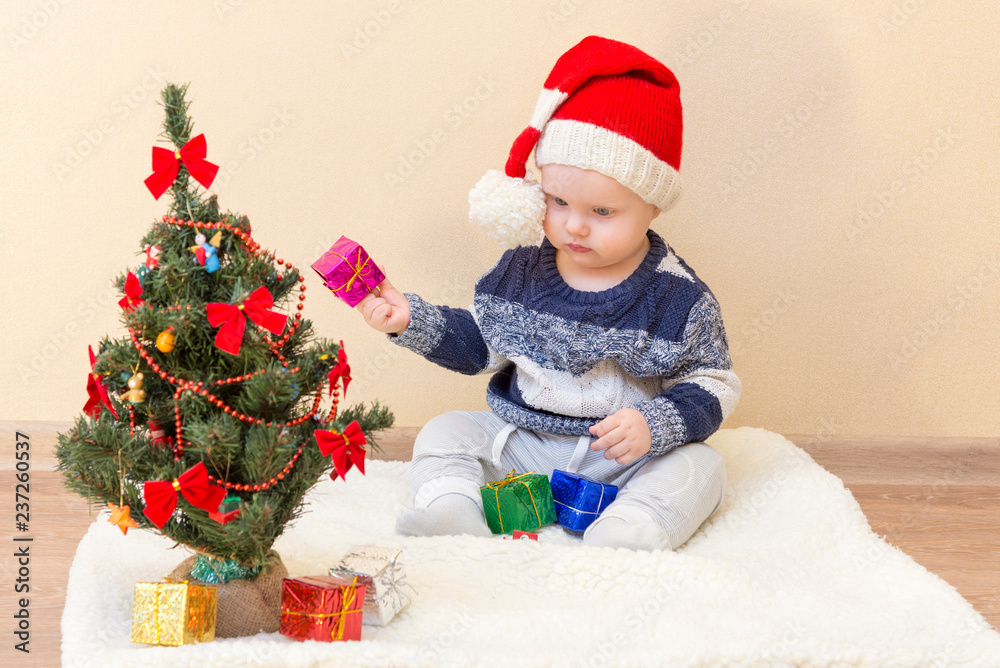 Funny baby in Santa Claus hat sitting near the Christmas tree with gifts