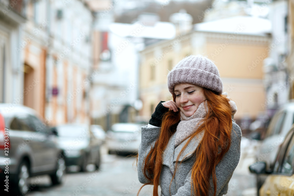 Pleased red haired girl wearing warm winter clothes walking down the street. Empty space