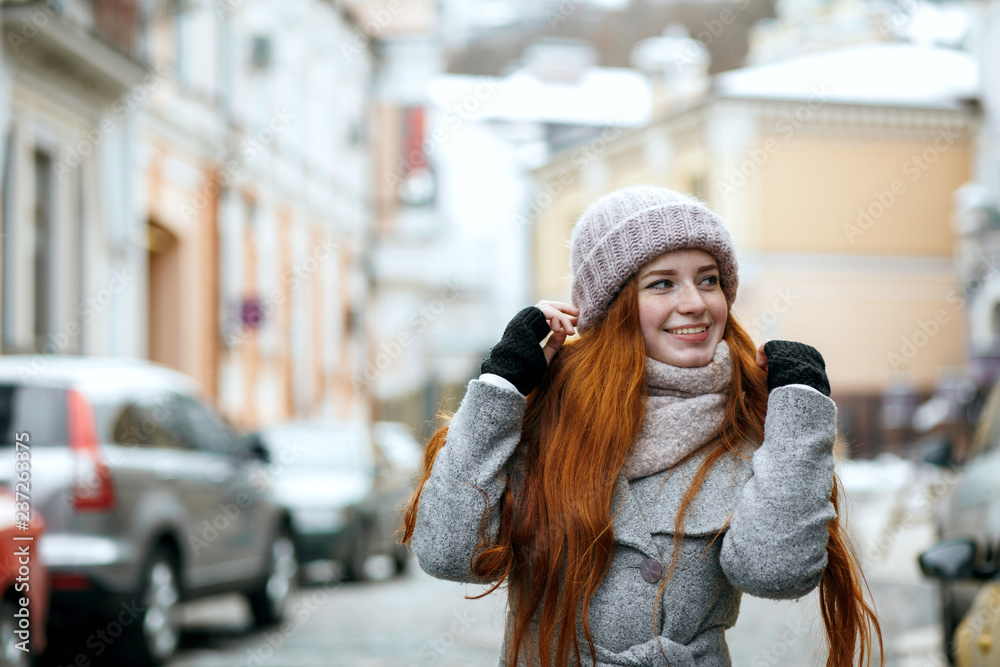 Cheerful red haired woman wearing warm winter clothes walking down the street. Empty space