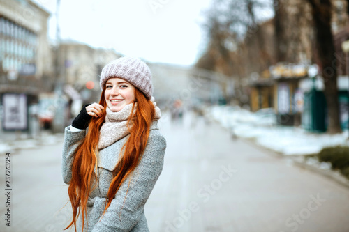 Urban portrait of positive ginger model with long hair wearing warm clothes walking at the city. Empty space