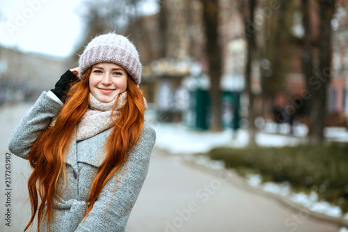 Urban portrait of happy redhead woman with long hair wearing warm clothes walking at the city. Empty space