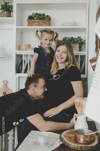 young family with pregnant woman and little girl sitting in scandinavian kitchen and having fun