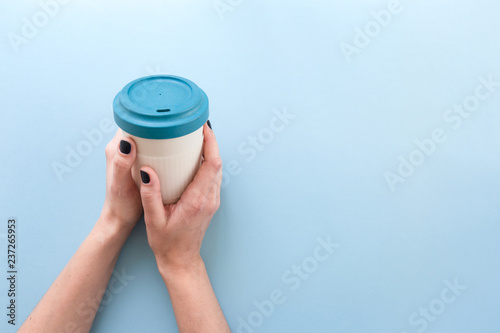 Hands holding bamboo reusable takeaway cup with lid on, on blue background.